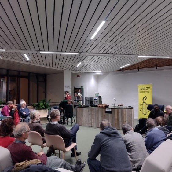 Een spreker voor een publiek op stoelen gezeten met een banner van Amnesty International vooraan